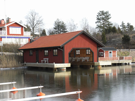 Rött båthus med trädäck längs fasaden och öppning mot vattnet.